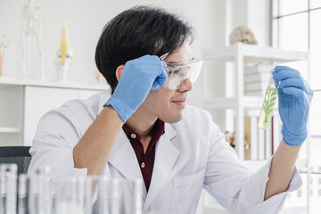 A male scientist with black hair wearing white coat and protective glassware holding a test tube with a green plant sample in a white laboratory or hospital.