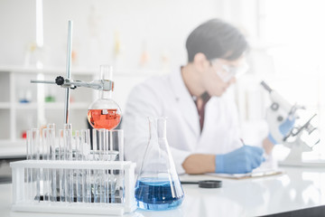 A male scientist with black hair wearing white coat and protective glassware writing and looking into a microscope in a laboratory setting with test tubes. Person blur in background.
