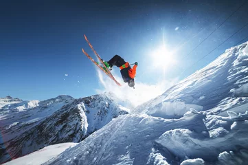 Deurstickers Low angle view athlete skier in an orange jacket does a back flip with flying powder of snow against a clear blue sky sun and snow-capped mountains of the Caucasus. © yanik88