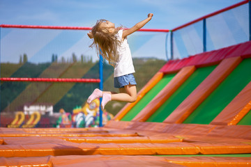 Fototapeta na wymiar Little pretty girl having fun outdoor. Jumping on trampoline in children zone. Happy girl jumping on the yellow trampoline in Amusement park