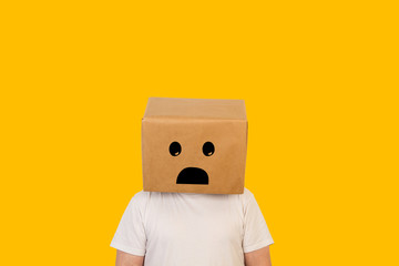Hombre con caja de cartón en la cabeza con un gesto de tristeza en la cara sobre fondo amarillo liso brillante. Vista de frente. Copy space - obrazy, fototapety, plakaty