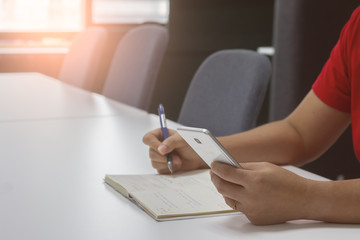 Close-up hand of a woman business owner wears a red casual hold smartphone and working in empty meeting room with blurred sunlight background. Social distancing and COVID-19 or work from home concept.