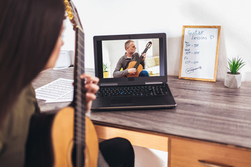 Focused girl playing acoustic guitar and watching online course on laptop while practicing at home....