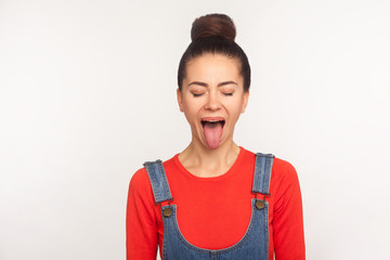 Portrait of funny stylish pretty girl with hair bun in denim overalls demonstrating tongue out...