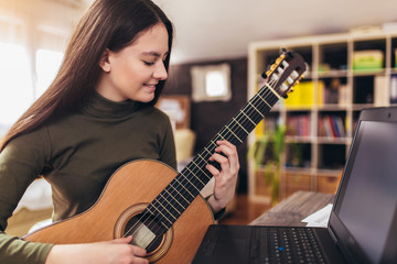 Fototapeta na wymiar Focused girl playing acoustic guitar and watching online course on laptop while practicing at home. Online training, online classes.