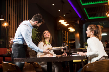 Smiling young female friends at a restaurant with waiter serving dinner