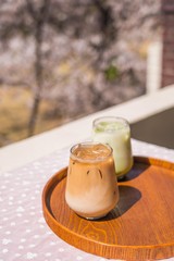Glass of Caffè Latte and Green Tea Latte on a wooden tray with pink cherry blossom tree background