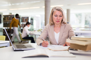 Mature confident woman with laptop and book in public library