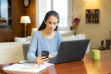 Young student girl looking at smartphone with laptop at home