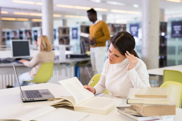 Thoughtful woman working with textbook and laptop in library