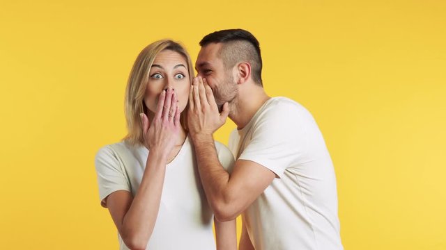 Young man whispering secrets to surprised woman over yellow background