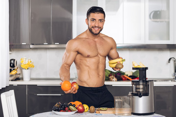 young fit man with fruits, in the kitchen, holding banana, orange, apple, kiwi. Preparing to make a fruit juice.