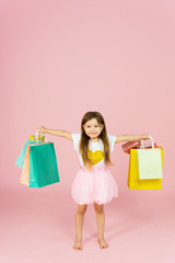 Little blonde girl enjoys her shopping on a pastel pink background with copyspace. Sale. Cute little girl with many multicolored shopping bags in studio