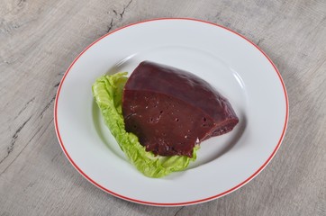 A piece of fresh beef liver on a white plate and decorated with a green lettuce leaf on a gray wooden background