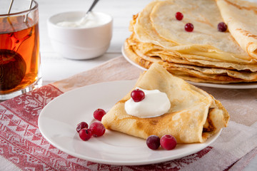  pancakes with berries and sour cream on a plate, tea on a towel with a red pattern on a white background,