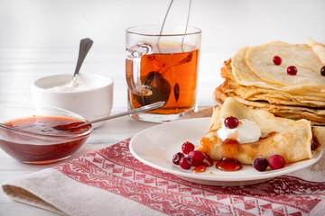 pancakes with berries and sour cream on a plate, tea, jam on a towel with a red pattern on a white background,
