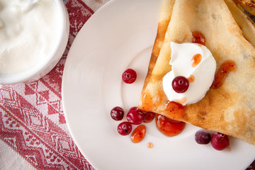 Russian pancakes with sour cream, cranberries and jam on a plate, tea, jam on a towel with a red pattern on a white background, top view,