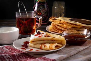 several pancakes with berries, sour cream and jam on a plate, a mug of black tea on a towel on a dark wooden