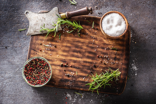 Top of view shot of square wooden cutting board and rustic cleaver on dark background with spices, fresh herbs and salt seasoning after meat was taken away. Food preparation items