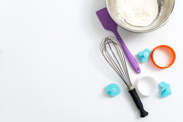 Kitchen items, spatula, whisk, baking dish, bowl of flour on a white background