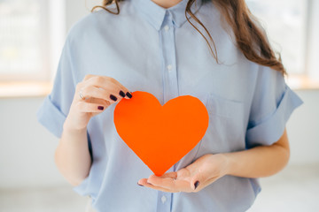 A young woman holds out a cut out construction paper heart near the window.