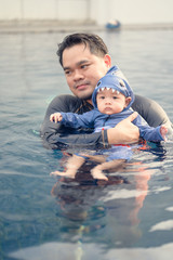 Fototapeta premium Asian father swimming with cute adorable baby in swimming pool. Smiling dad and little child, newborn son having fun together.