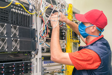 A technician in a medical mask switches wires in the server room. A man in a red cap works in a...