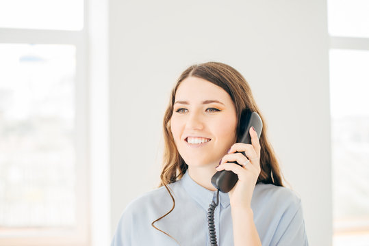 beautiful brunette talking on telephone and smiling. close up of young girl holding receiver and cord near the window