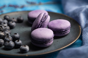 Fototapeta na wymiar Purple macarons or macaroons cakes with blueberries on ceramic plate on a blue concrete background. Side view, selective focus.