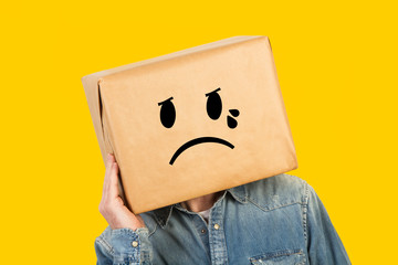 Hombre con caja de cartón en la cabeza con gesto de tristeza y con lágrimas en los ojos sobre fondo amarillo liso brillante aislado. Vista de frente. Copy space - obrazy, fototapety, plakaty