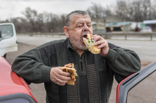 Portrait of Caucasian senior driver being delighted while gobbling lyulya kebab in lavash near his car