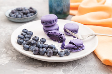 Purple macarons or macaroons cakes with blueberries on white ceramic plate on a gray concrete background. Side view, selective focus.