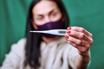 a brunette girl wearing a purple medical mask shows a white electronic thermometer into the camera. green background. covid-19. protect the health. hygiene and quarantine.