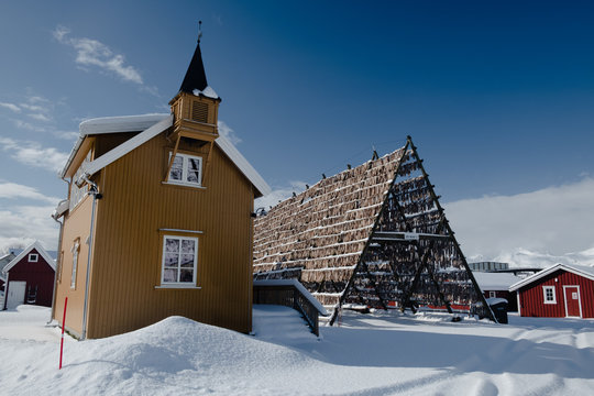 Yellow house in scandinavian style with a belfry. Drying rack is fully hanged by famous stockfish. Winter photo shows symbols of faith and fishery which are very important for Norwegian culture.