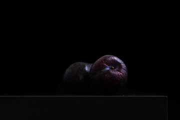 PAIR OF PLUMS ON BLACK TABLE AND BLACK BACKGROUND. DARK FOOD. PICTORIAL. STILL LIFE