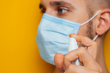 Portrait of a young guy in a mask on a yellow background. Disinfects hands with an antiseptic. Sprays the pest control. The concept of coronavirus. Protection against germs and viruses.