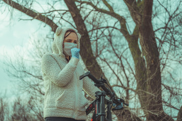 Young woman in medical mask and gloves coughing, holding on to rudder of bicycle in countryside. Female protecting yourself from diseases on walk. Concept of threat of coronavirus epidemic infection.