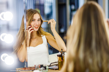 Young beautiful girl doing makeup in front of mirror and talking on mobile phone.