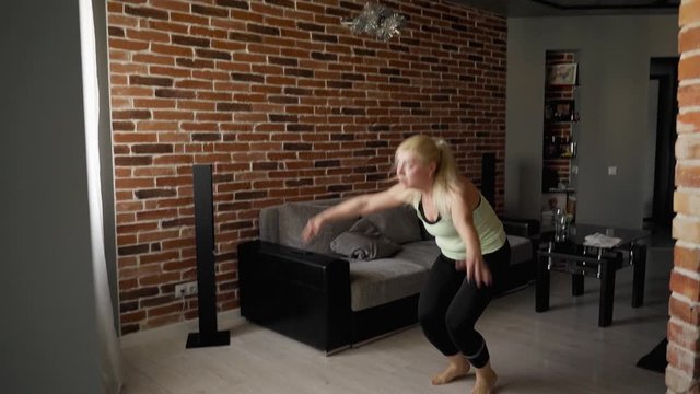 Trainings at home: A strong beautiful fitness girl makes burpee exercise. She make Jump to the bar, push-ups, jump up. Woman begin workout at her living room. She in athletic sport wear. Slow motion