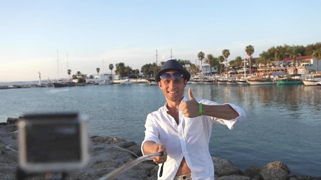 Vacation Man making a selfie photo or video when he showing a thumbs-up Hand gesture of himself using a selfie-stick and action camera in the marine bay. Cheerful traveling time concept footage.