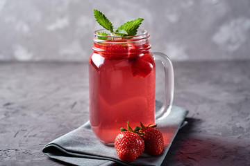 Homemade strawberry tea with mint in mason jar on gray concrete table background, copy space. Cold summer berry drink in sparkling glasses. Fresh vitamin beverage