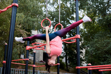 Fototapeta na wymiar Young strong brunette woman, wearing pink top and blue leggings, hanging on gymnastic rings, doing twine. Slim fit sportswoman, doing stretching fitness exercises. Healthy lifestyle concept.
