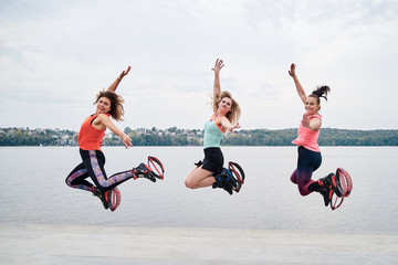 A group of young fit slim women in kangoo jumps, jumping in the air in front of city lake in summer. Three girls, wearing colorful sports outfit, doing exercises. Healthy lifestyle concept.