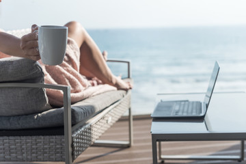 Obraz premium Beautiful brunette woman freelancer wrapped in a plaid resting on a couch and sunbathing on the terrace with a cup of coffee at summer day. Sea view. WFH. Working from home