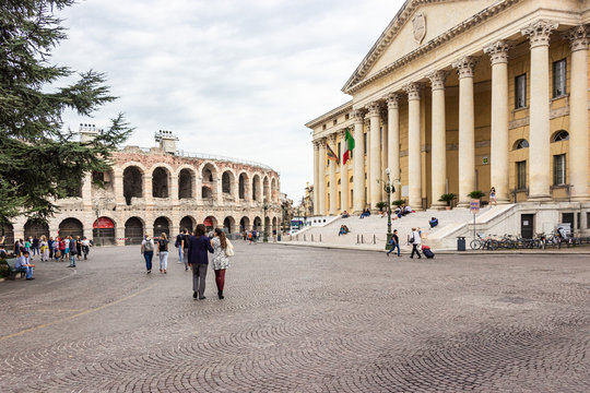 Tourists walking around the Piazza Bra square on a cloudy day, inspect the Arena and building of Palazzo Barbieri Comune di Verona and take pictures against its background in Verona, Italy.