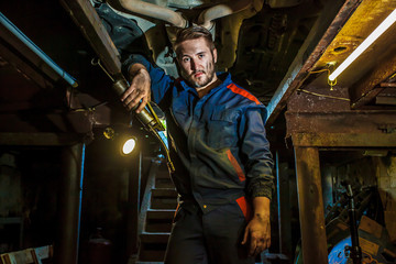 Obraz na płótnie Canvas Portrait of a mechanic standing in a protective blue suit in a garage under a car. Car service concept. Car repair and maintenance. Mechanic examining under the car at the repair garage.