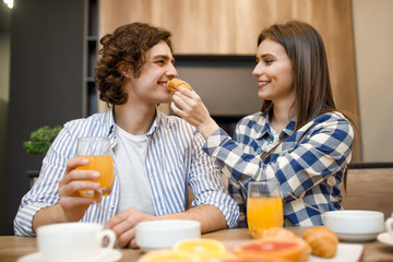 Portrait of pretty couple feeding each other during breakfast in kitchen, cornflakes with milk, orange and croissant at the table