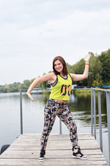 Young brunette sportswoman, wearing yellow top with zumba sign and colorful leggings on wooden pier. Full-length portrait of Zumba fitness instructor, dancing by city lake. Healthy lifestyle concept.