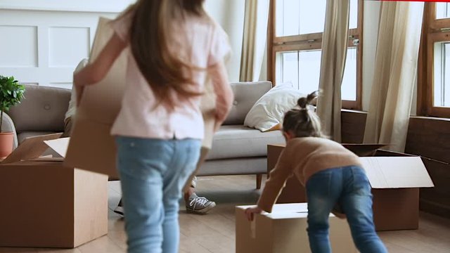 Happy young married couple sitting on couch, laughing at funny video on digital tablet while small children siblings running playing with huge cardboard boxes in renovated new living room, moving day.