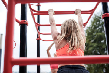 Fototapeta premium Young women, wearing pink tops and dark leggings, hanging on horizontal bars outside by city lake. Slim sportswomen, doing fitness exercises. Healthy lifestyle concept. Strength training.